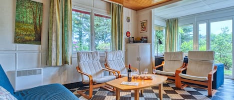 A cozy room with a wooden ceiling, a checkered floor, a sofa, two armchairs, a small table with a bottle and glasses, and large windows with curtains.