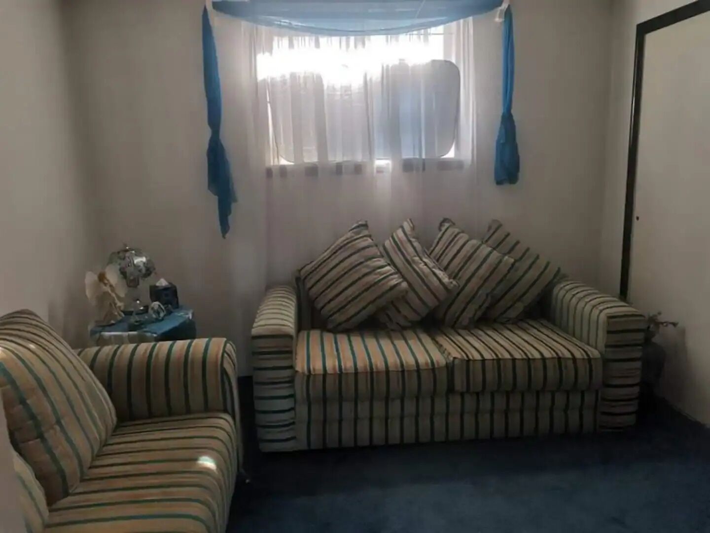 A corner room with a striped sofa and armchair, a small table with a lamp, and a window with curtains.