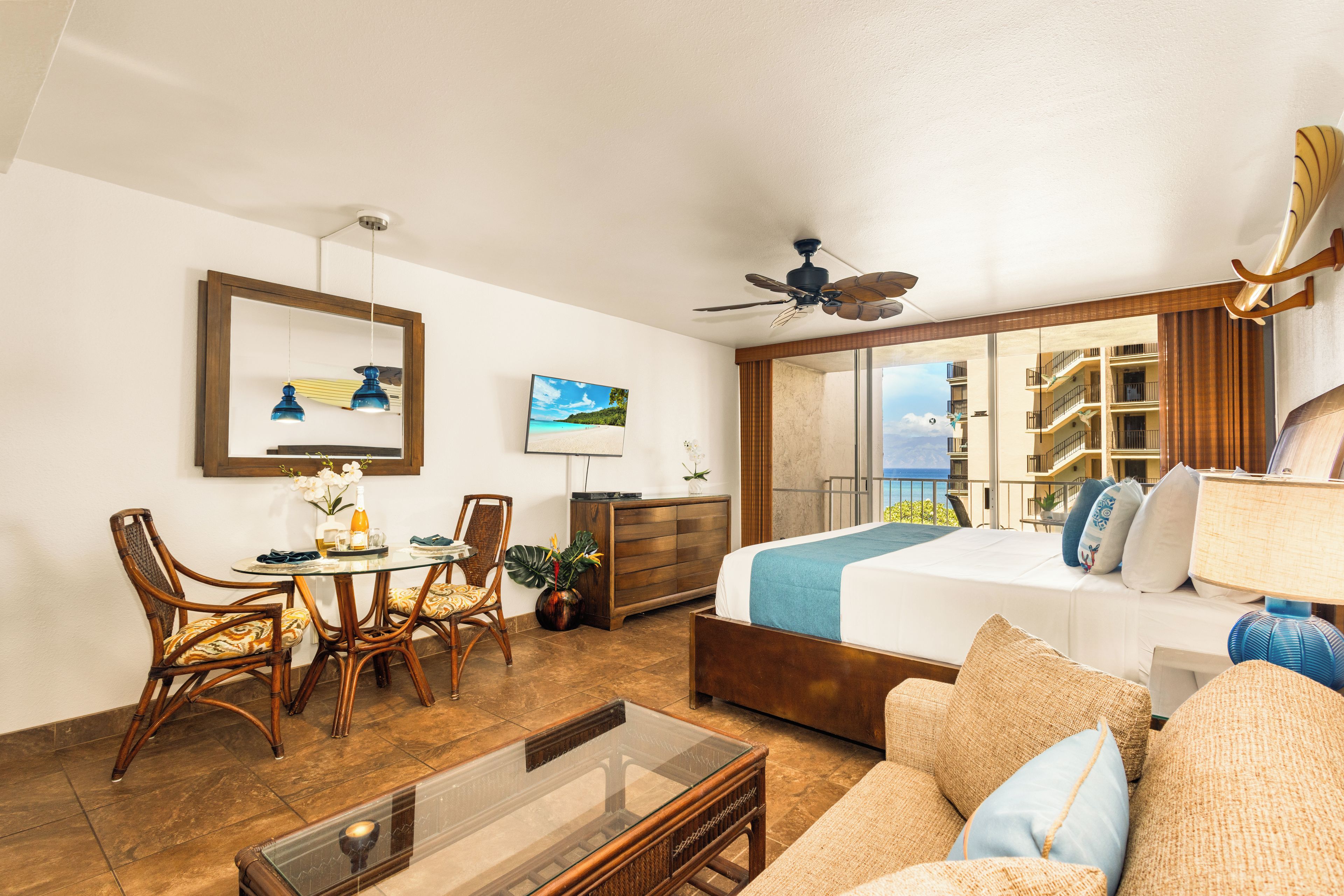 Habitación de hotel con cama, zona de estar con mesa y sillas, ventilador de techo y ventana grande con vista a un edificio y al cielo.