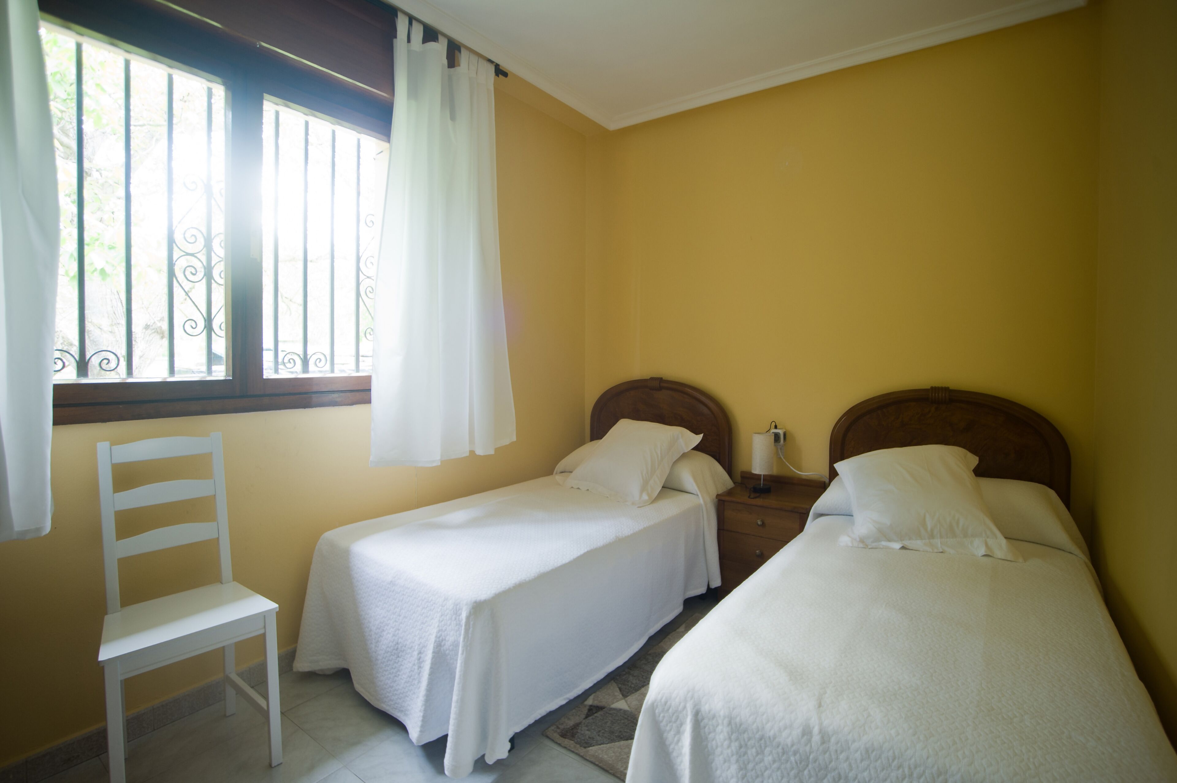 A room with two single beds, a wooden chair, a nightstand with a lamp, and a window with curtains.