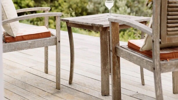 Una terraza de madera con dos sillas y una mesa, un vaso de agua sobre la mesa, y un fondo de vegetación densa.