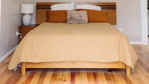 A bedroom with a wooden bed, a nightstand, a lamp, and a hanging light fixture. - Wainui Stream Cottage (Raglan)
