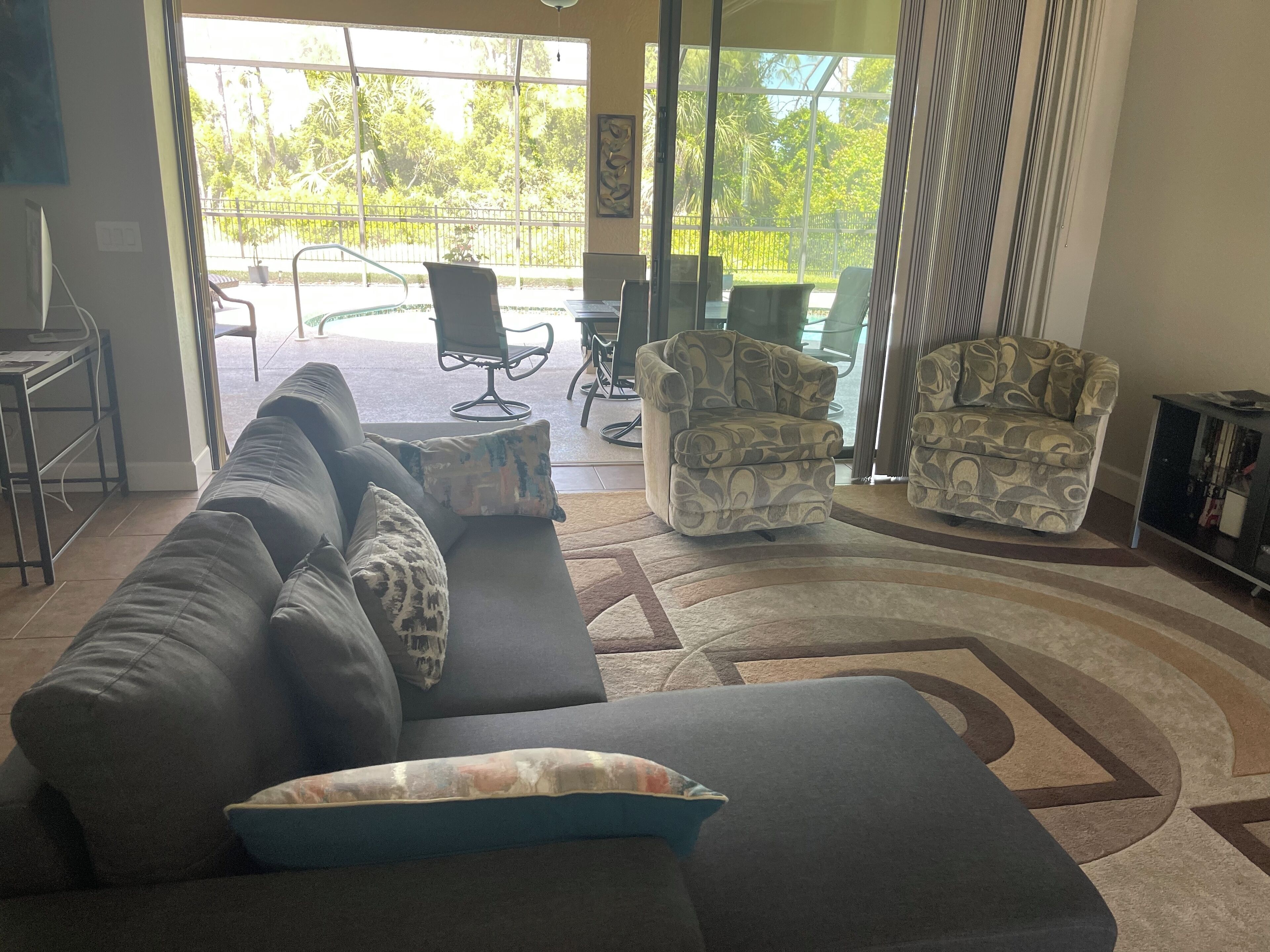 Una sala de estar con un sofá, sillones y una mesa de centro. Hay un ventanal que da a un área exterior con un patio y sillas.