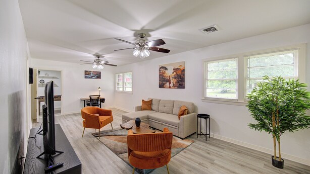 A modern living room with a sectional sofa, a coffee table, and a television on a wooden stand. There are two ceiling fans, a potted plant, and a large window with blinds. - Spacious house with massage chair and game room (Beaumont)