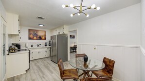 A modern kitchen with white cabinets, a stainless steel refrigerator, and a dining area with a glass table and leather chairs. - Spacious house with massage chair and game room (Beaumont)