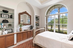 A bedroom with a large arched window, a wooden vanity with shelves, and a bed with a white comforter. - This waterfront home on Marco Island is the perfect vacation destination for families or groups. (Marco Island)