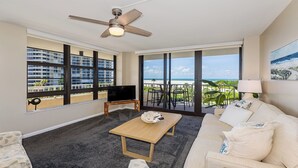 A spacious living room with a large TV, a ceiling fan, and a view of the beach through large windows.