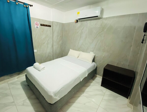 A hotel room with a bed, a blue curtain, a no smoking sign, an air conditioning unit, and a small black cabinet.