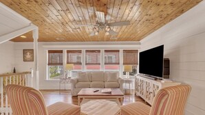 Een ruime woonkamer met houten plafond, een groot raam met zonwering, een beige bank, gestreepte fauteuils, een salontafel en een televisie op een kast.