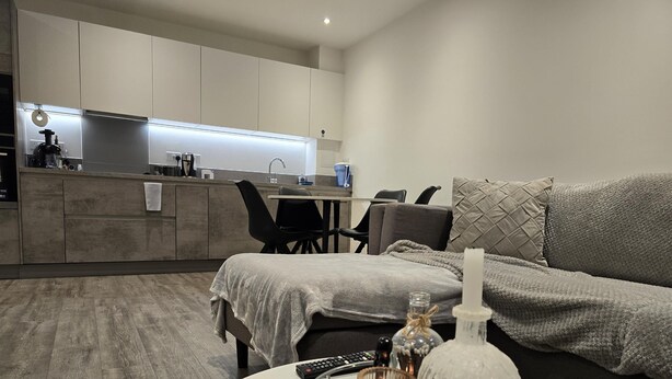 Un salon moderne avec un canapé gris, une table basse sur laquelle se trouvent une bougie et une bouteille, et une cuisine en arrière-plan.
