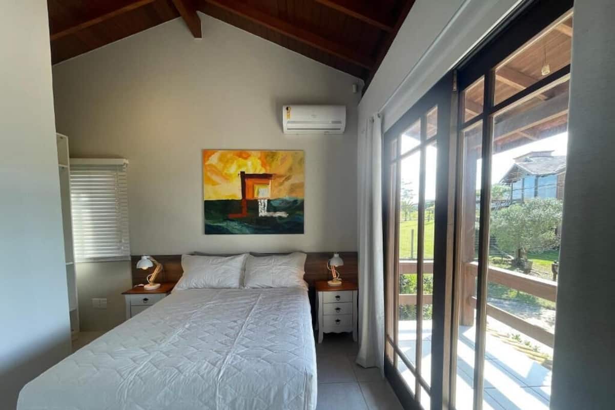 A bedroom with a large bed, a painting on the wall, and a view of the outdoors through large windows.