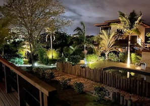 Vue nocturne d’un complexe hôtelier doté d’une piscine, d’une terrasse en bois et d’un aménagement paysager de style tropical.