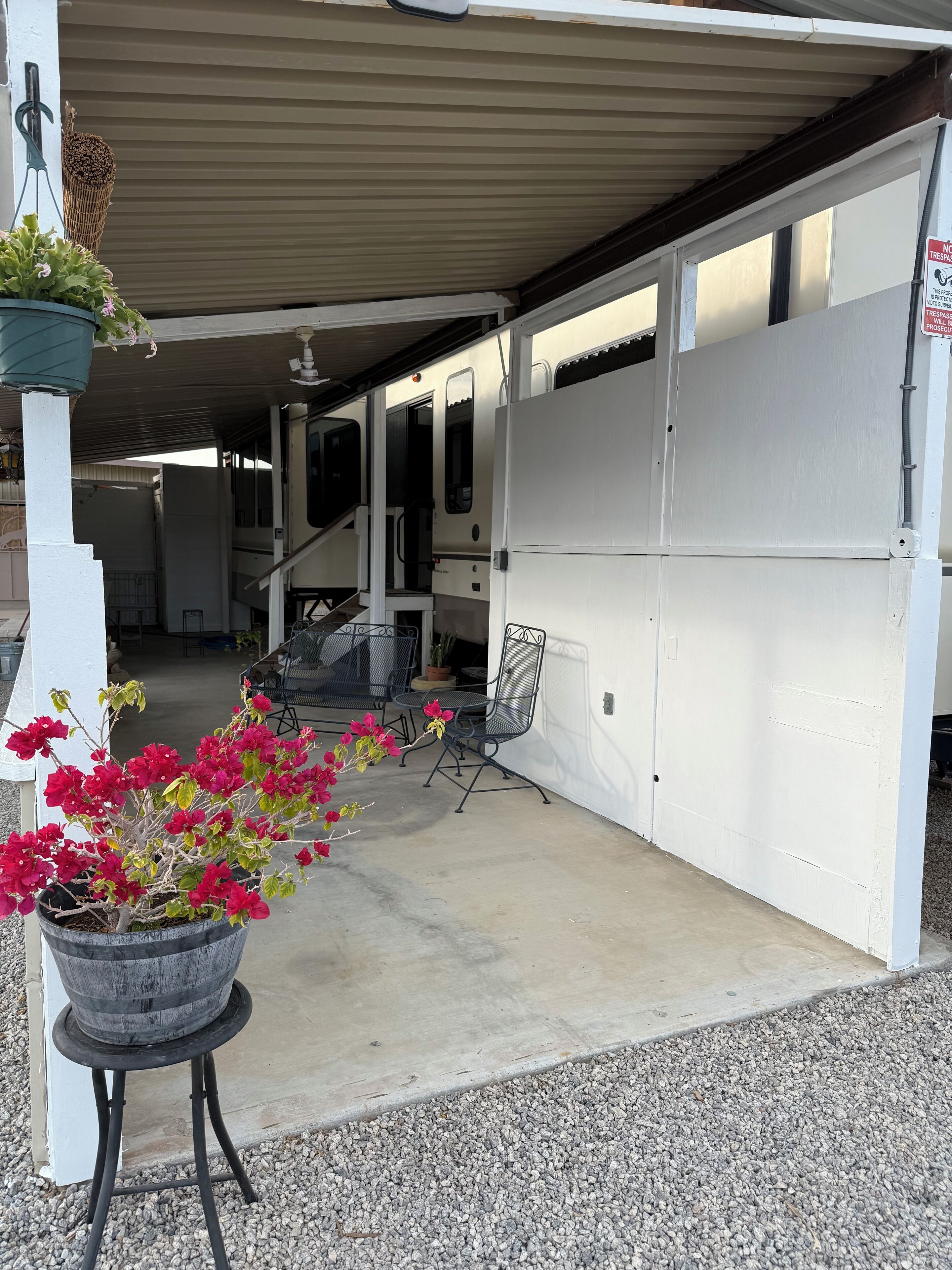 A covered outdoor area with a concrete floor, a metal table and chairs, and a potted plant with red flowers.