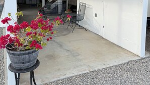 Un espace extérieur abrité, doté d’un sol en béton, d’une table et de chaises en métal, ainsi que d’un pot de fleurs contenant des plantes aux fleurs rouges.