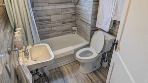Un baño moderno con bañera, inodoro y lavamanos. Destaca un diseño de pared de piedra y un ventanal que proporciona luz natural.