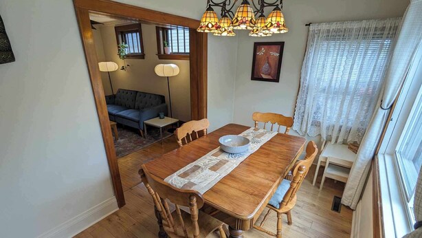 Comedor con mesa de madera, sillas y lámpara de araña. Se observa un sofá y una ventana con cortinas al fondo.