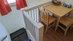 A small dining area with a wooden table and chairs, a white railing, and a window with red curtains.