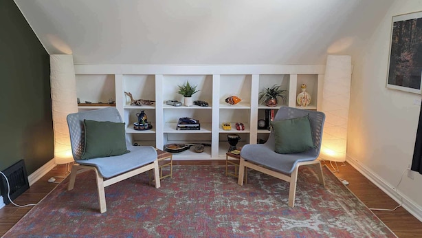A cozy living room with two chairs, a patterned rug, and a white shelving unit with various decorative items.