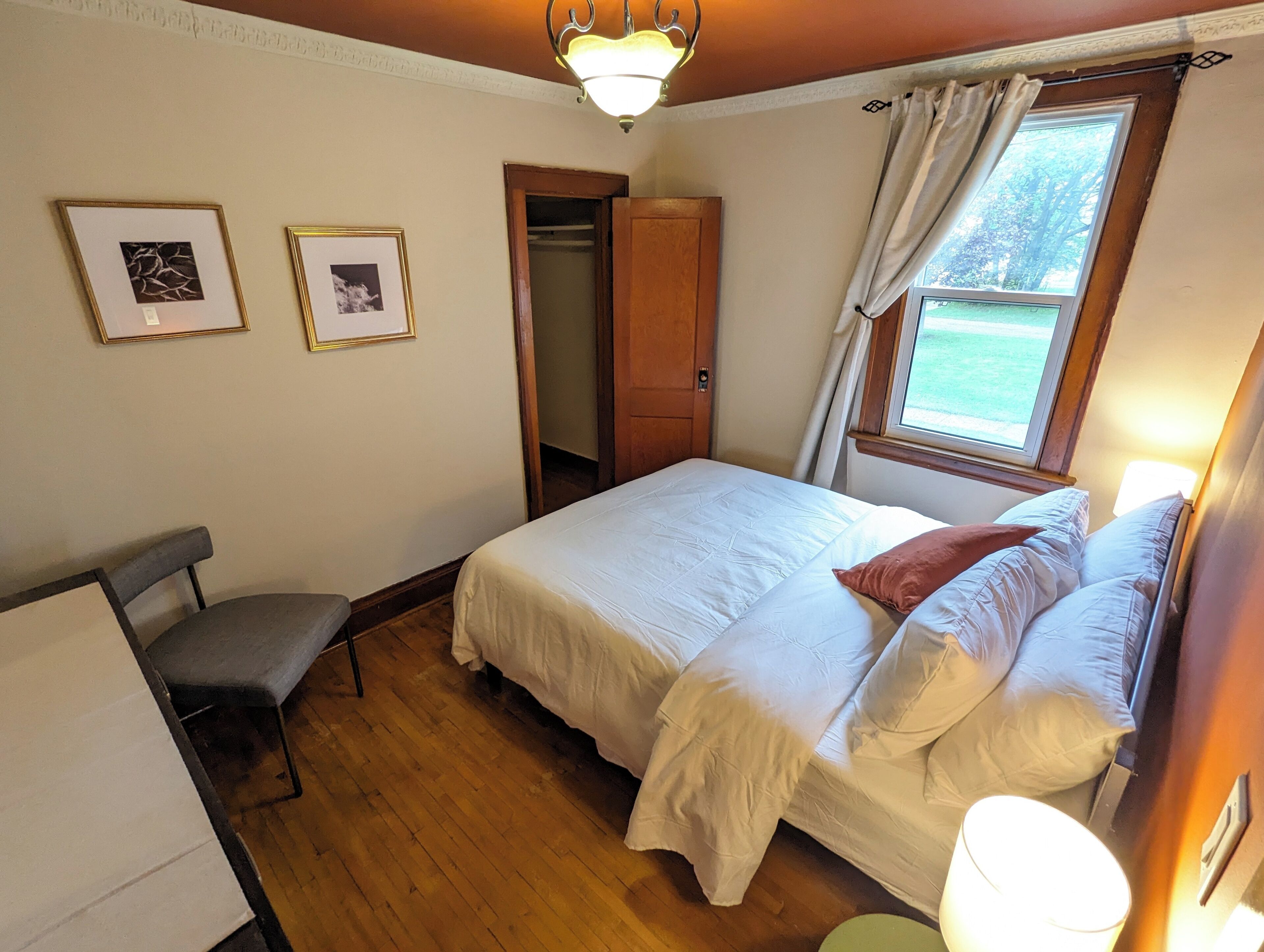 A bedroom with a bed, a chair, a desk, a window with curtains, and framed pictures on the wall.
