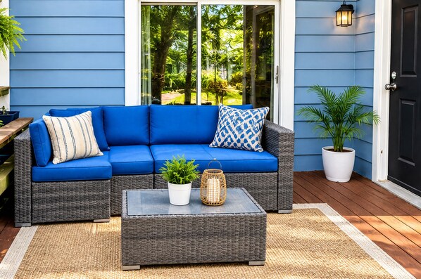 A blue sofa with cushions, a wicker coffee table, and potted plants on a wooden deck.