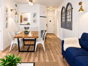 Een eethoek met een houten tafel en witte stoelen, een blauwe bank en een raam met decoratieve luiken.