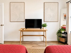 Ein Zimmer mit Schreibtisch aus Holz, einem Flachbildfernseher, zwei gerahmten abstrakten Kunstwerken an der Wand, einer Kübelpflanze, einem Korbwägelchen und einer roten Bettdecke.