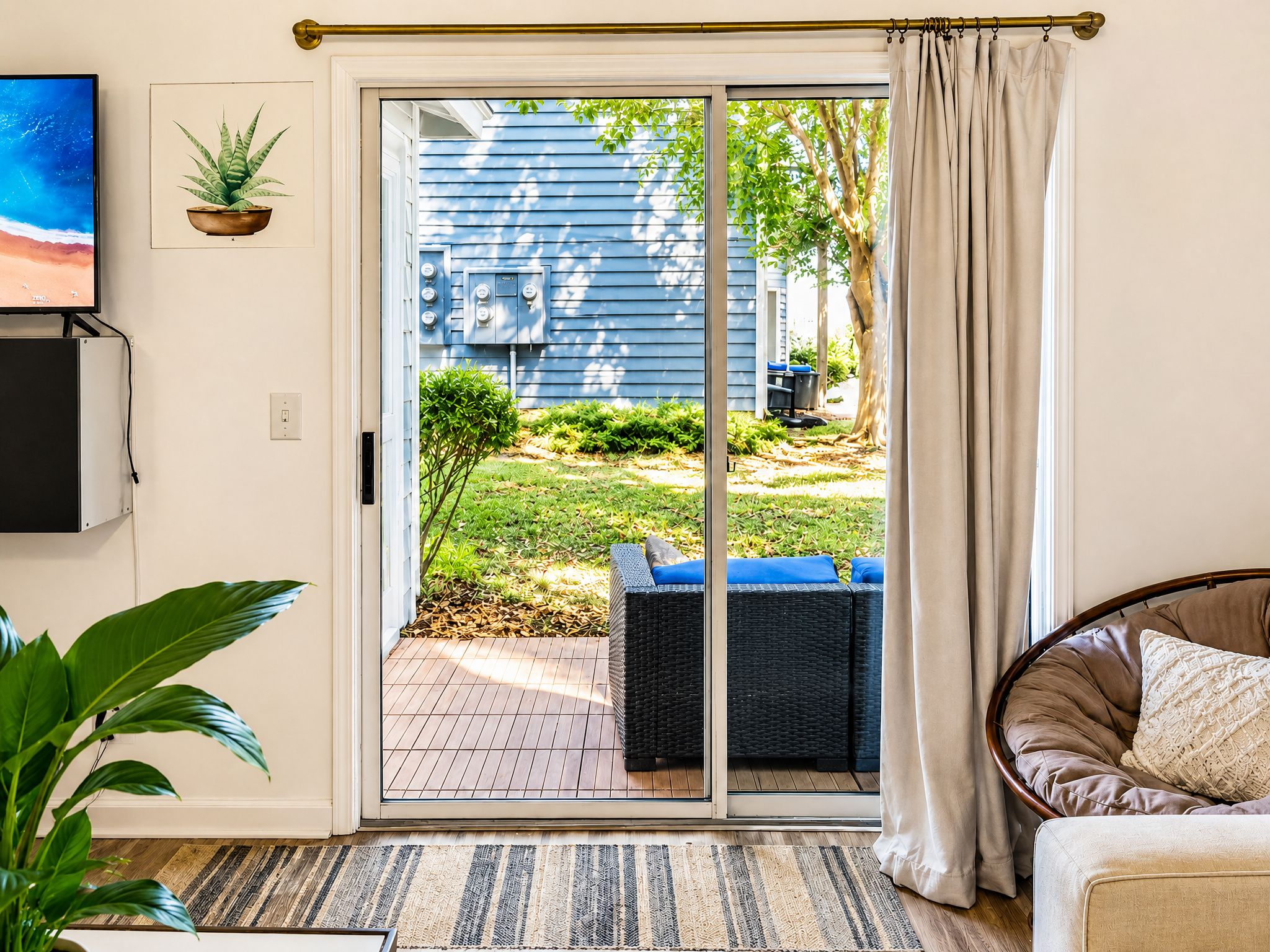 A room with a sliding glass door leading to a backyard, a television mounted on the wall, and a wicker chair with cushions.