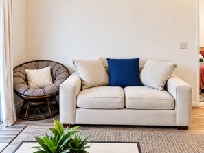 Ein Wohnzimmer mit einem beigen Sofa, einem Korbstuhl und einem blauen Kissen.