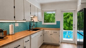 Una cucina con mobili bianchi, un piano di lavoro in legno, una finestra vista piscina e una porta che dà su un balcone.