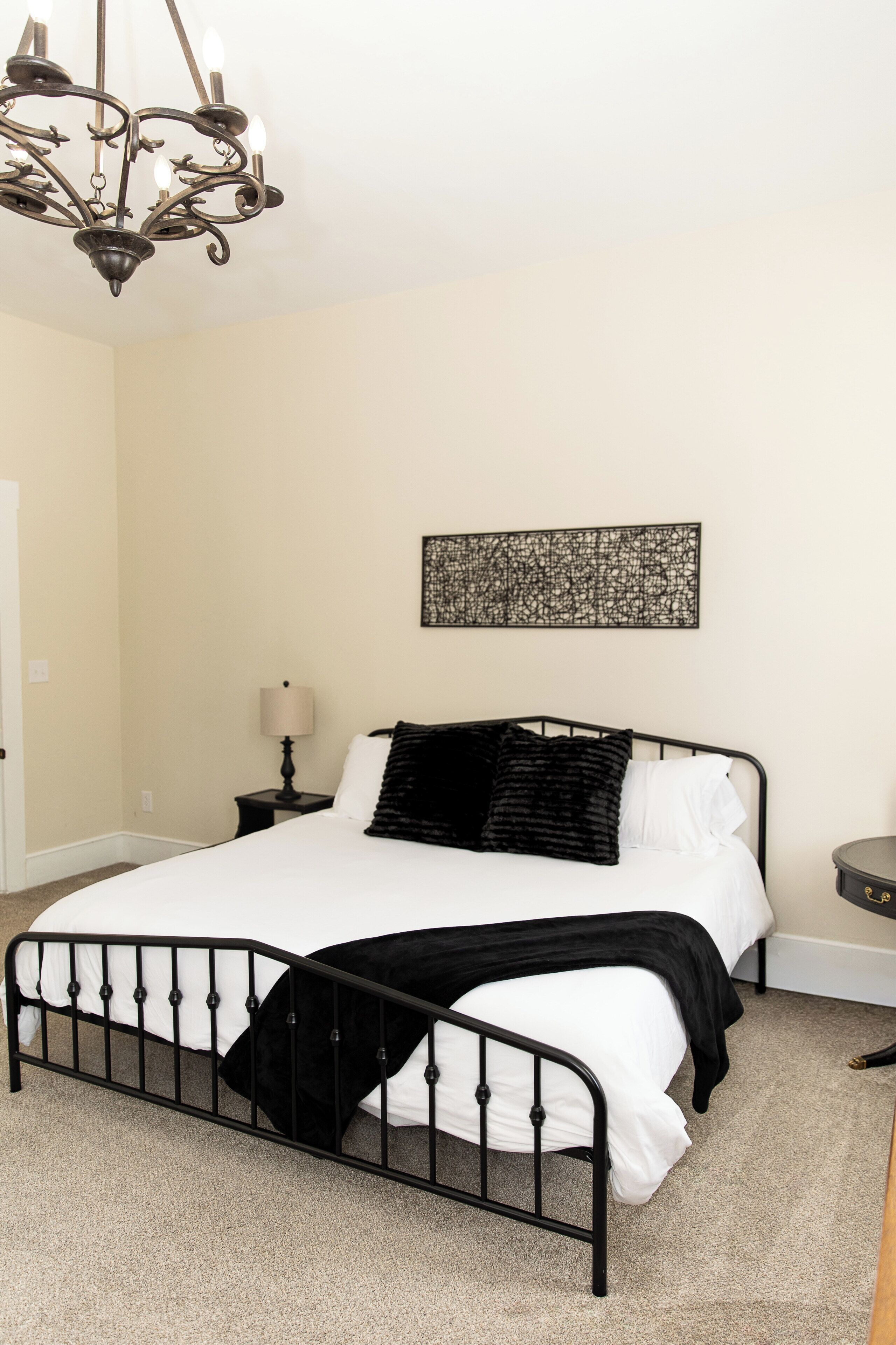 A bedroom with a bed, a chandelier, a nightstand, a lamp, and a wall art piece.