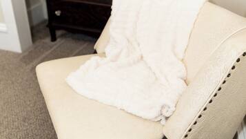 Una poltrona beige con una coperta bianca, un comò in legno scuro e un secchio nero in una stanza con pavimento rivestito di moquette.