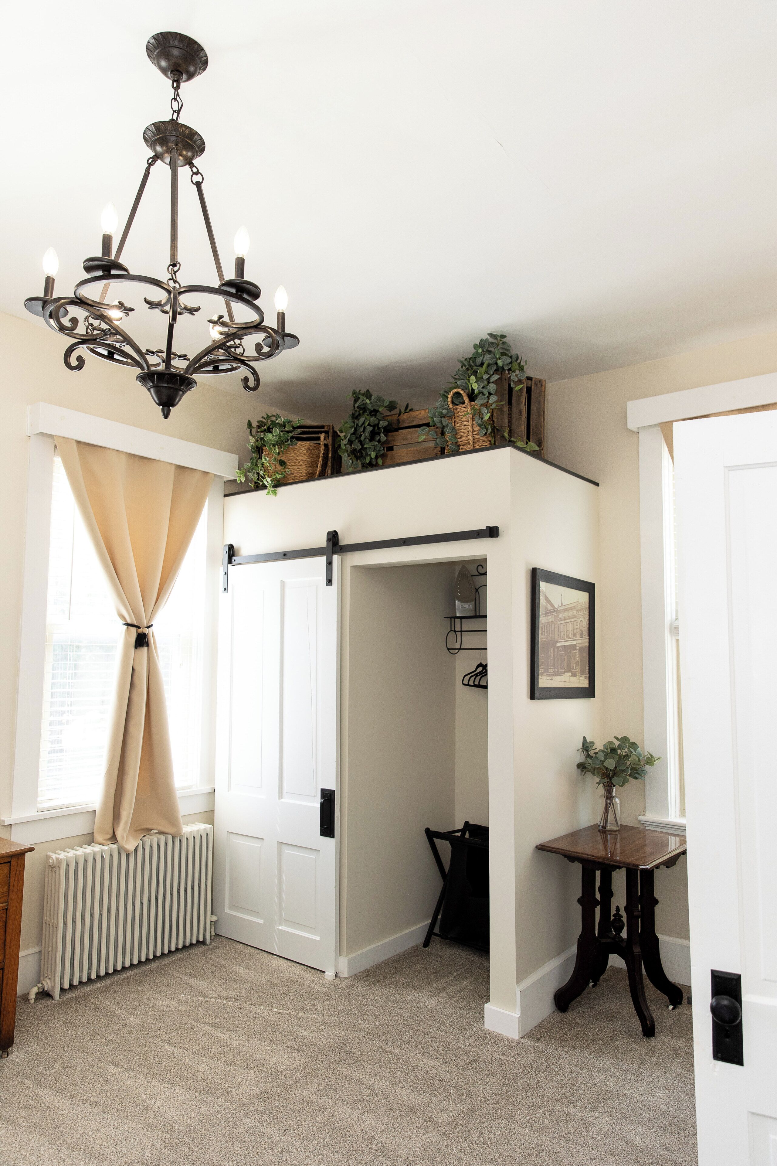 A room with a chandelier, a radiator, a door with a black handle, a small table with a vase, and a framed picture on the wall.
