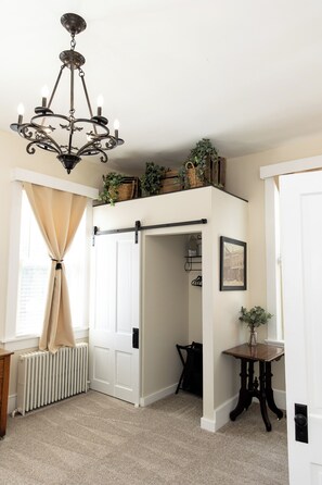 Una habitación con candelabro, radiador, puerta con pomo negro, una pequeña mesa con un jarrón y un cuadro enmarcado en la pared.