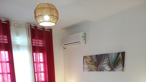 Un lit bien fait, avec du linge de lit blanc, une tête de lit en bois et un tapis rouge qui apporte une touche de couleur. Un climatiseur mural est fixé au mur, et une œuvre d’art décorative est accrochée au-dessus du lit.