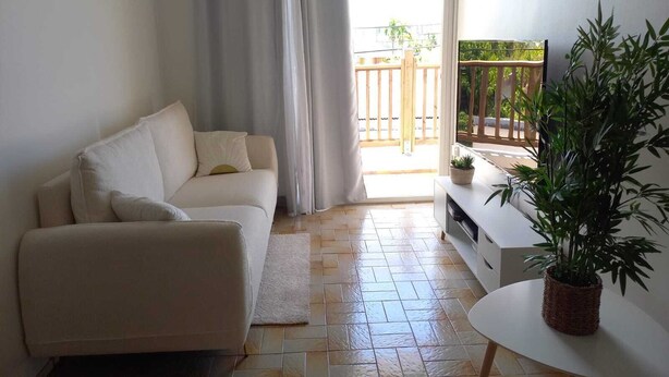 Un salon avec un canapé beige, une petite table blanche et une plante en pot. Une porte-fenêtre coulissante donne sur un balcon doté d’une rambarde en bois et offrant une vue sur la verdure.
