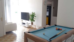 Une table de billard avec des billes, un téléviseur, une plante en pot et un canapé blanc dans une pièce dont le sol est recouvert de carreaux.