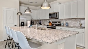 Una cocina moderna con armarios blancos, encimeras de granito, electrodomésticos de acero inoxidable e iluminación colgante.