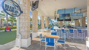 Un área de bar al aire libre con pilares de piedra, sillas azules y blancas, y una gran pantalla de televisión.