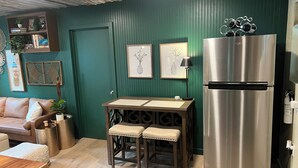 A compact kitchen with a wooden table, two stools, a stainless steel refrigerator, and a green wall with decorative items. - Cozy farmhouse cabin near Kilgore and Longview (Longview)