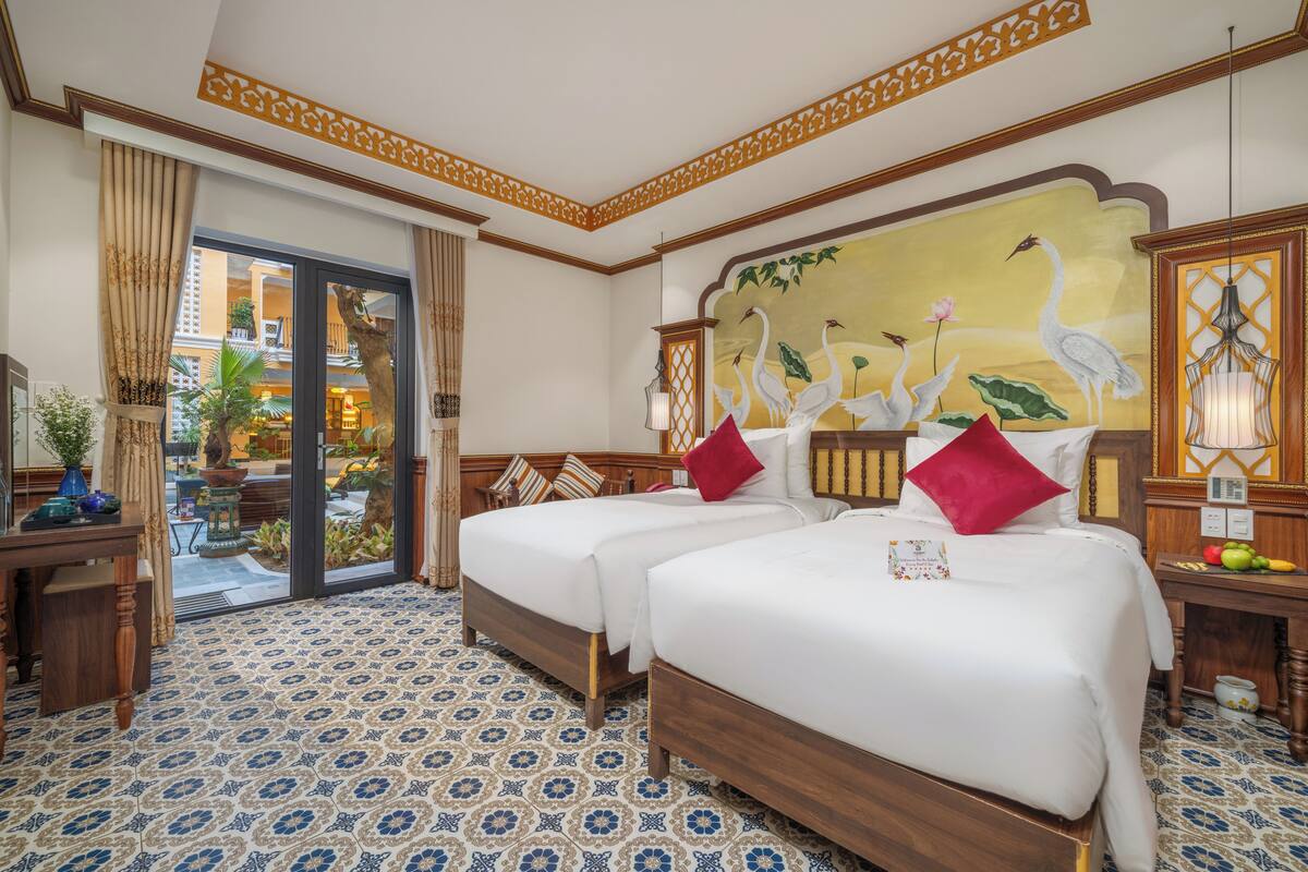 A hotel room with two beds, a large mural of birds, a dining area with a table and chairs, and a view of the outdoor garden.