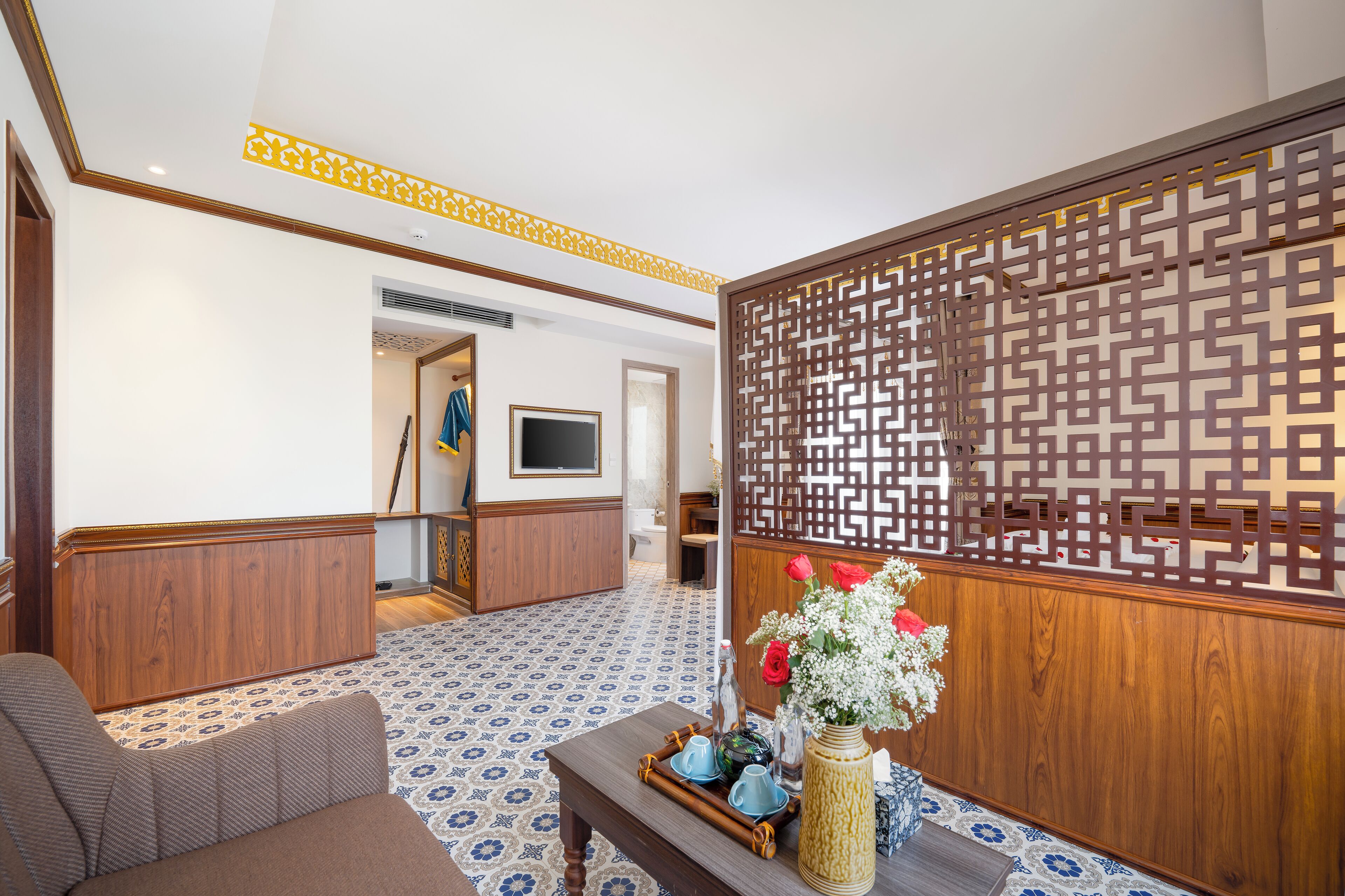 Un vestíbulo de hotel con suelo estampado, un separador de madera, un sofá, una mesa de centro con objetos decorativos y un televisor al fondo.