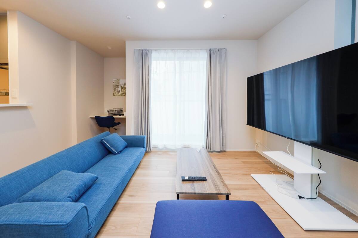 A modern living room with a blue sofa, a wooden coffee table, a flat-screen TV, and a large window with curtains.