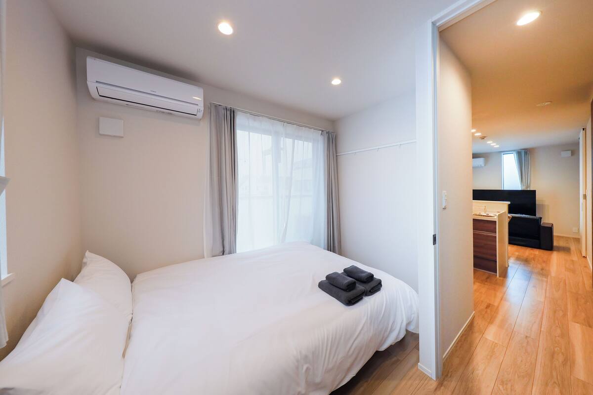 A hotel room with a bed, white walls, an air conditioning unit, a window with curtains, and a wooden floor.