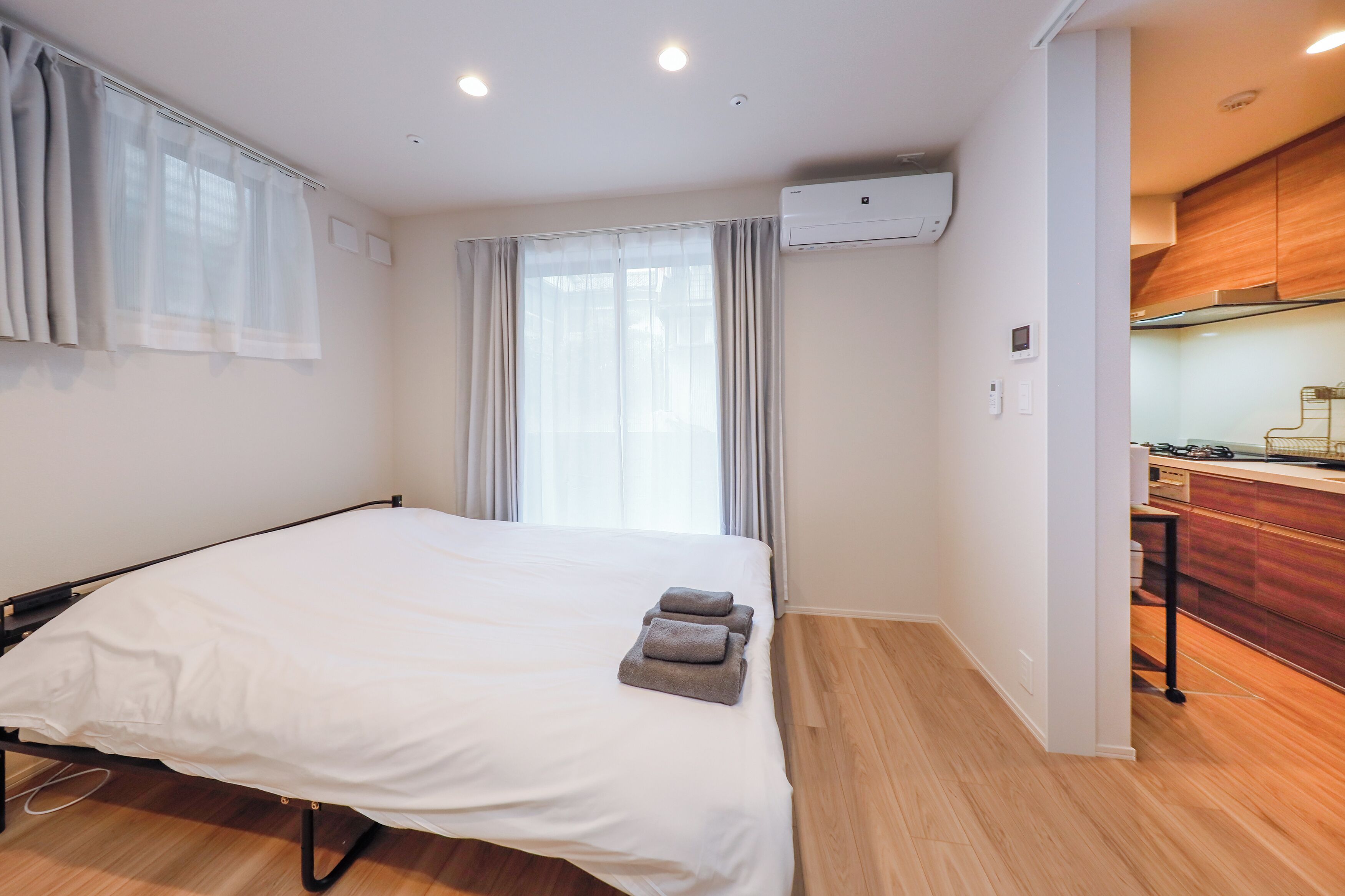 A bedroom with a bed, a window with curtains, an air conditioning unit, and a kitchenette.
