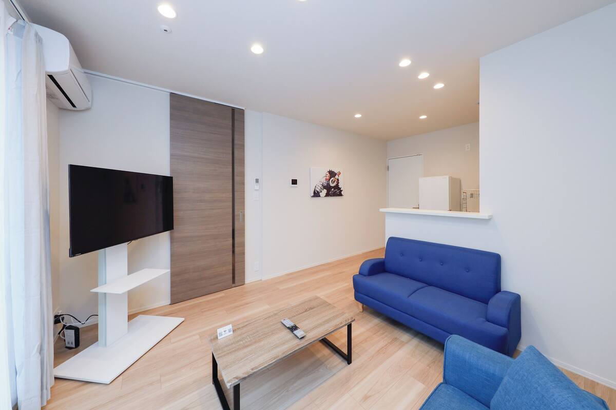 A modern living room with a flat-screen TV, a blue sofa, a wooden coffee table, and a white air conditioning unit.