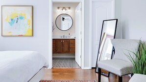 Makuuhuoneessa on sänky, tuoli, kehystetty taulu ja ikkunasta avautuu näkymä kylpyhuoneeseen, jossa on peili ja pesuallas.