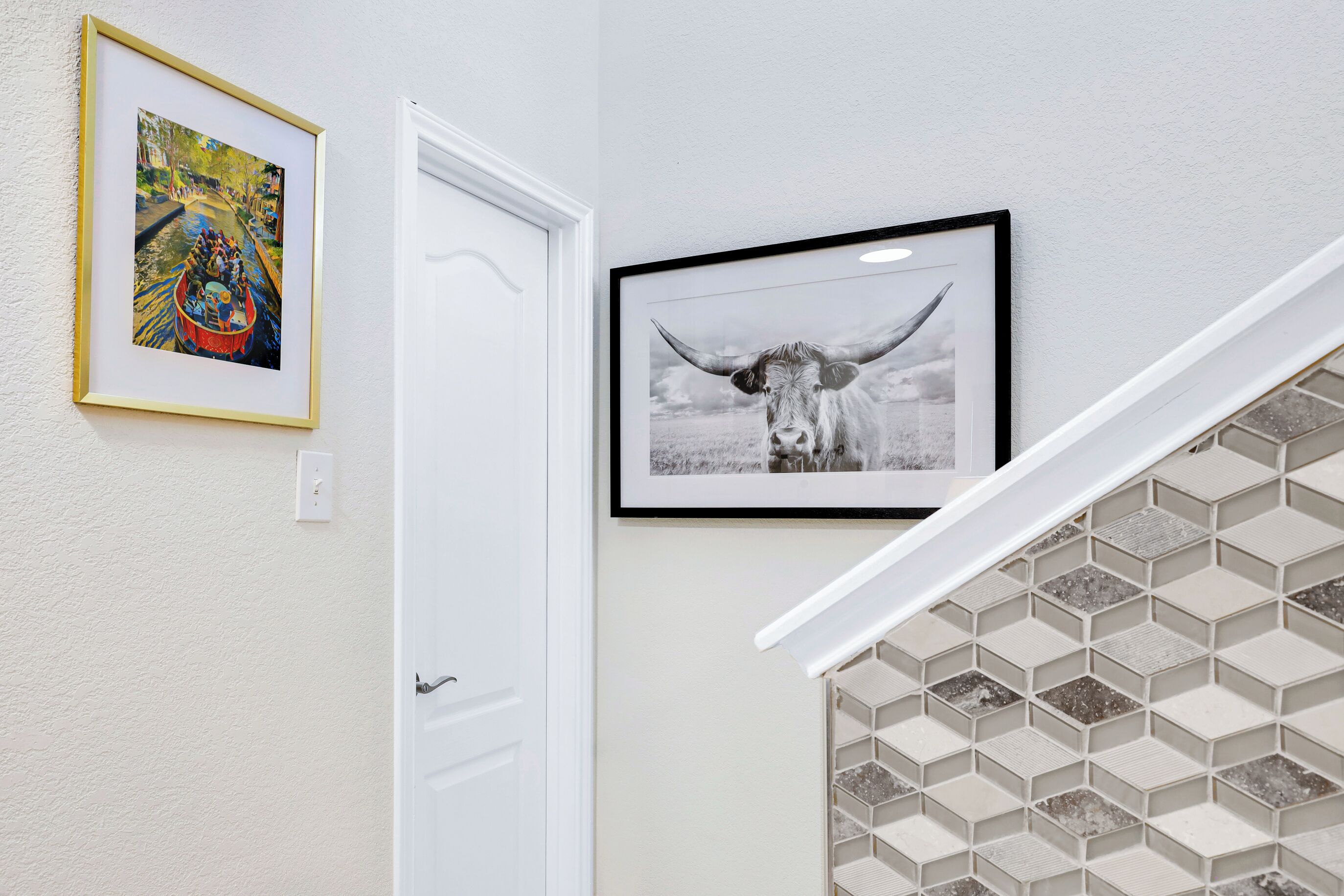 Un pasillo con una puerta blanca, una fotografía en blanco y negro de una vaca enmarcada, y un cuadro colorido.