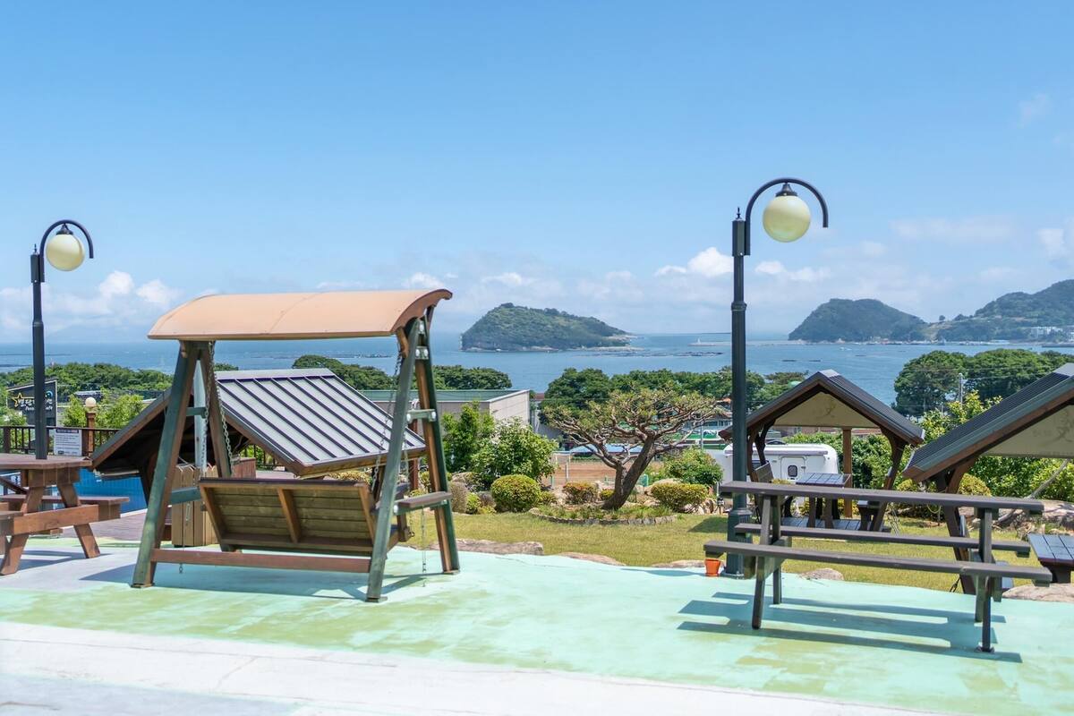 A playground with swings and a picnic table, overlooking a scenic bay with mountains and a clear sky.