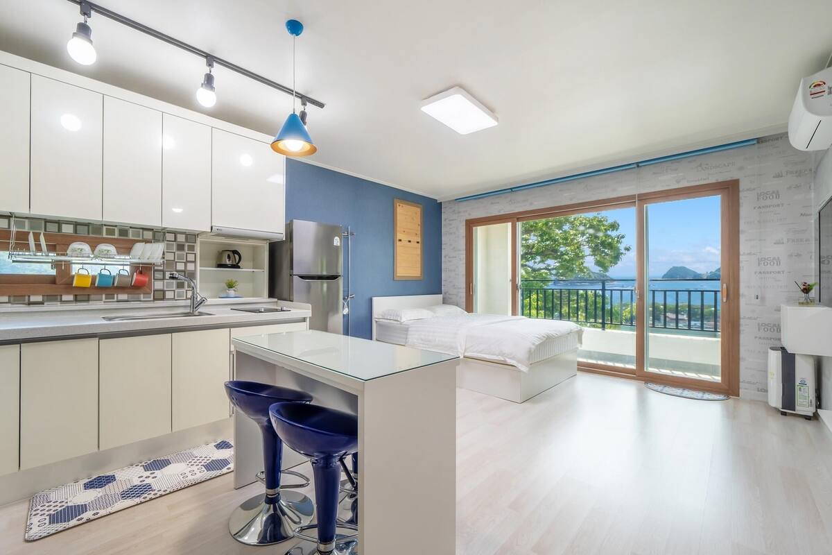 A modern kitchen with white cabinets, a central island, and blue bar stools. There is a bedroom with a bed and a balcony with a view of the sea.