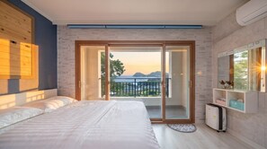 A hotel room with a bed, a small desk, a wardrobe, and a balcony with a view of the sea. - Namhae Familyae Pension (Namhae)